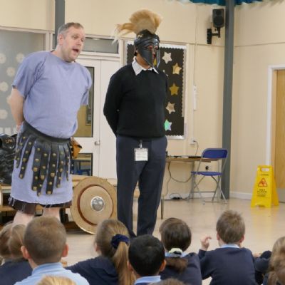 Ancient Greek presenter and our teacher in a helmet the pupils are in the foreground.